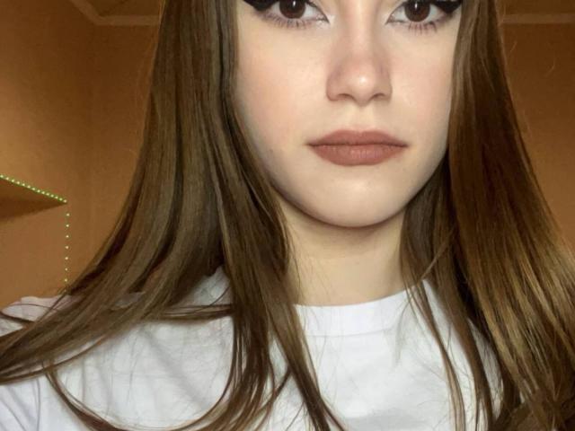EstellVanderiet's face, white shirt, brown hair, thick eyeliner, full lips, pale skin, looking straight at camera, soft light, slight shadow under chin.
