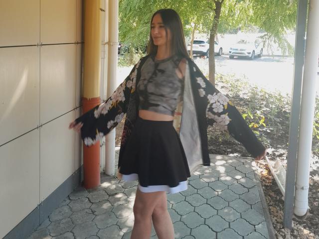 Omella with long black hair, wearing a black floral kimono, gray tie-dye crop top, and black skirt, standing in a sunlit outdoor area, smiling.