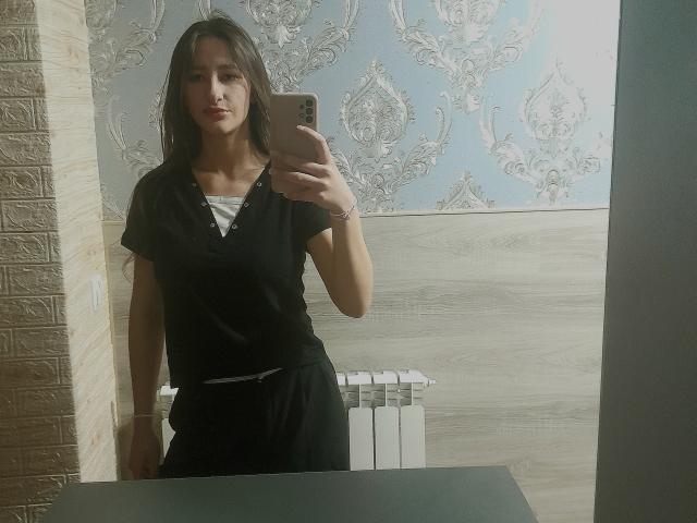 Omella with medium tits in a black shirt and black pants, taking a mirror selfie with a serious expression, holding a rose gold iPhone, standing in front of a radiator.