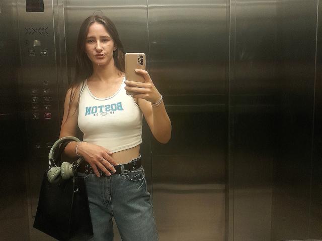 Omella with long brown hair, white crop top, "HOTON" text, blue jeans, holding phone, black bag, green jacket, in metallic elevator, standing, smiling, medium tits, slim.