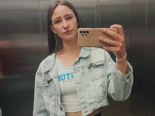Omella with long brown hair, light skin, wearing a denim jacket over a white crop top, holding a gold iPhone, taking a selfie in a mirror.