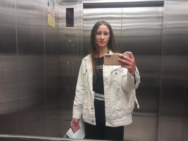 Omella with long brown hair, wearing a white jacket and black shirt, holding a phone and a ticket, standing in a metallic elevator, smiling at the camera.