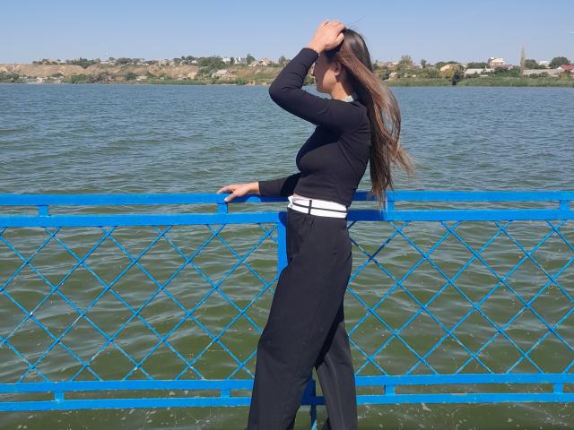 Omella with long brown hair, black long-sleeve top, black pants, white belt, standing by blue metal fence, hand in hair, looking at water. Omella's ass is slightly visible.