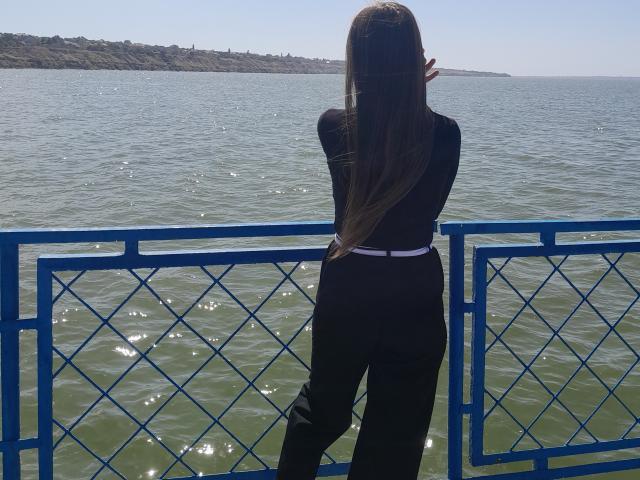 Omella with long brown hair, black long-sleeve top, and black pants, stands with her back to the camera, looking at a blue-railed, water-filled view. Ass is visible.
