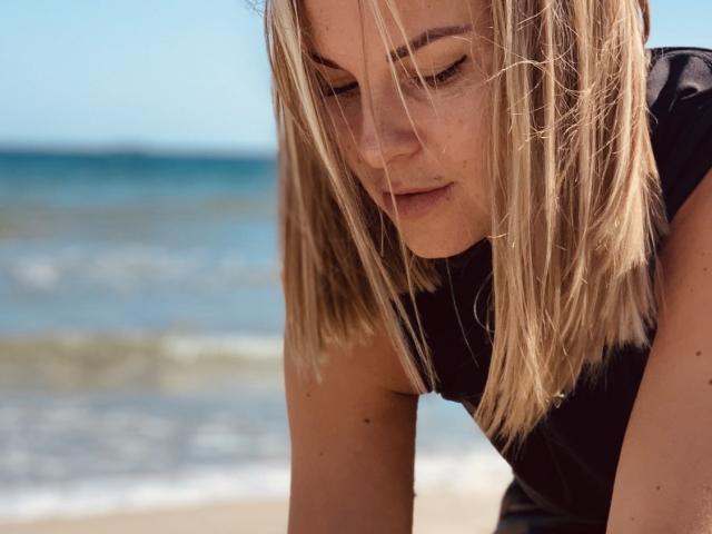 BelleLisaG with blonde, shoulder-length hair leans forward, eyes down, in a black sleeveless top. Her fair skin and light freckles are visible. She has subtle makeup with a focus on her eyes. The photograph has a natural, candid feel with a slight blur on the background, emphasizing her relaxed, contemplative posture. The image captures a moment of stillness and introspection.