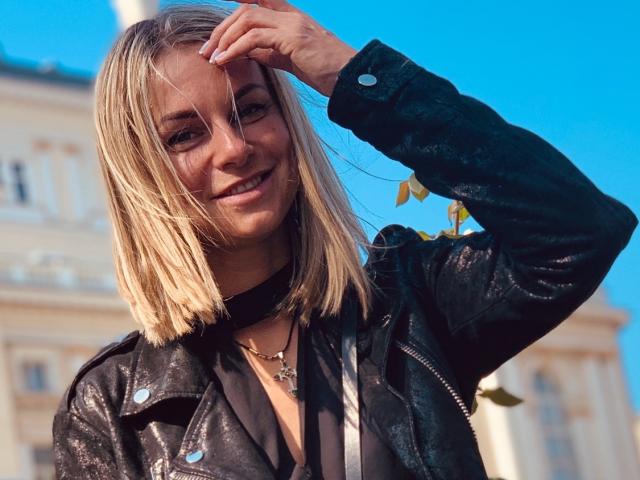 BelleLisaG has a medium build, with blonde shoulder-length hair, slightly parted to reveal her face. She's wearing a black leather jacket with metallic studs, a black choker, and a thin necklace with a small pendant. Her right hand is raised, fingers touching her hair, with a subtle smile on her face. The bright blue sky forms the background, and a blurred, beige building is partially visible. Her hair has a straight, flat texture. The image uses natural light, highlighting the shiny texture of her leather jacket. She has a light skin tone.