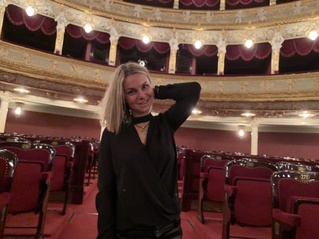 BelleLisaG stands in a grand, opulent theater with ornate, gold-accented upper balconies and maroon-upholstered seats. She has shoulder-length blonde hair, a slight smile, and wears a black long-sleeve top with a choker necklace. Her right hand is raised, resting on her head. The room is lit by small, round lights, and the red carpet matches the seats. The composition focuses on BelleLisaG in the foreground.