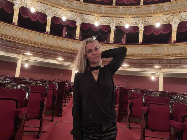 BelleLisaG with blonde hair, black top, and black pants, standing in an ornate theater with red chairs, hand behind head, small skull necklace, slight smirk, looking at camera.