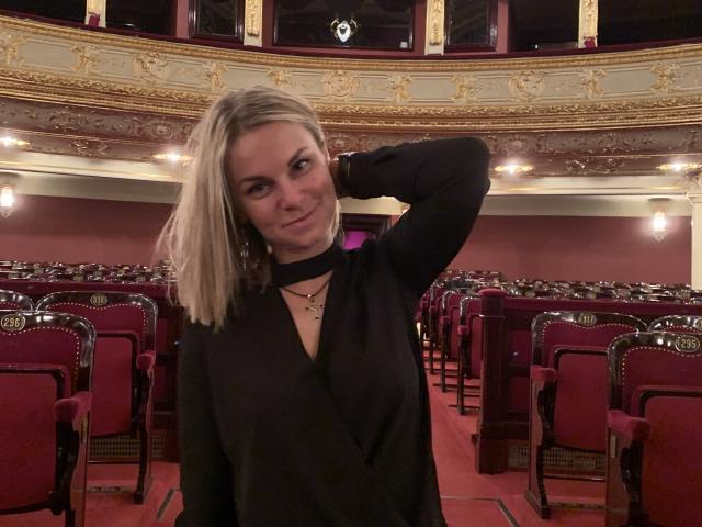 BelleLisaG stands in a grand, opulent theater, wearing a black, long-sleeved top with a deep V-neck. She has blonde hair, slightly wavy, and wears a necklace with a small cross. Her right arm is raised, hand resting on her head. The theater is filled with red, upholstered chairs, and the walls are adorned with gold and white decorative elements. Soft, warm lighting illuminates the space. Her expression is slightly smirking, with a relaxed, confident demeanor.