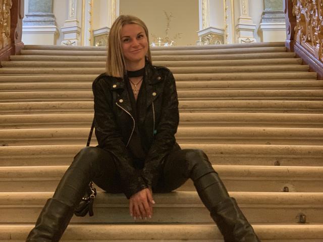 BelleLisaG sits on a wide, beige staircase with her legs spread apart. She has straight, shoulder-length blonde hair and a slight smile. She wears a black leather jacket with gold buttons, a black choker, and black leather pants with knee patches. Her right hand rests on the step, and her left hand is on her knee. The staircase has ornate wooden railings on either side and a luxurious, ornate background. The composition is balanced, with BelleLisaG centered. The lighting is warm.