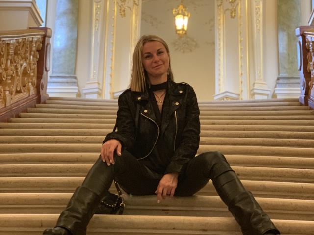 BelleLisaG sits on a grand staircase, legs spread apart, in a luxurious, ornate hallway. She wears a black leather jacket, black pants, and knee-high boots. Her blonde hair is straight, falling to her shoulders. She has a slight smile, with a necklace visible. The background includes a golden chandelier, decorative white and gold walls, and a carved wooden railing on the left. The image has a warm, slightly blurry quality, emphasizing the opulent setting. Her confident posture contrasts with the elegant surroundings.