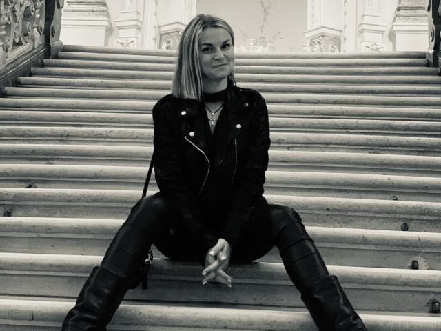 BelleLisaG sits on a wide, stone staircase, legs spread, hands clasped. She's a blonde woman with a bob haircut, wearing a black leather jacket, dark pants, and knee-high boots. Her confident smile contrasts with the monochrome, textured steps. The image uses a high-contrast, black-and-white style, emphasizing her sharp, edgy look. Her posture is relaxed yet assertive. The background shows ornate, classical architecture. The composition centers on BelleLisaG, highlighting her as the focal point.
