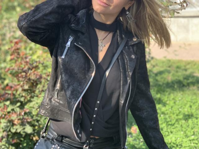 BelleLisaG stands outdoors, wearing a black leather jacket with a textured pattern, a black choker, and a black blouse. She has light brown hair, slightly wavy, partially covering her face. She's also wearing black pants with a belt and a necklace with a small pendant. The jacket has a zipper and a small pocket on the left side. The image is sharp with a green, leafy background. The light is natural, highlighting the textures of the jacket and her hair. Her right hand is raised, touching her hair.