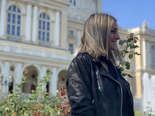 BelleLisaG, blonde, holds a small green leafy branch in her right hand, wears a black leather jacket. She's in the foreground, slightly right, with a blurred, classical European building in the background. Her jacket has a shiny texture, and she's looking to the right with a subtle smile. Bright blue sky above. Her hair is straight and medium-length. Photo has a natural, outdoor light.