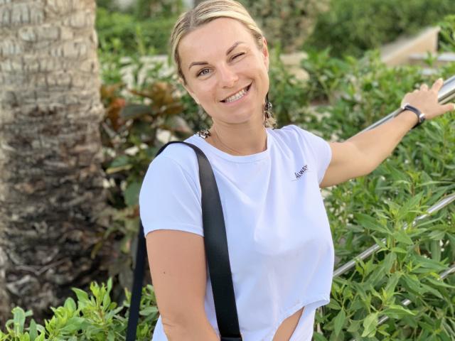 BelleLisaG stands outdoors with a palm tree on the left. She has blonde hair in a low bun, fair skin, and a winking smile. She wears a white athletic tee with "Aeropostale" on the chest, and a black strap crosses her chest. Green shrubs fill the background. She holds a metal railing with her right hand, wearing a black wristband. The image is bright and clear.