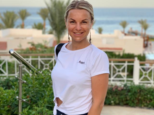 BelleLisaG stands in a sunny outdoor setting with a clear blue sky and ocean in the background. She has light brown hair in a simple, casual style, and is smiling at the camera. She wears a white, short-sleeved, athletic t-shirt with a small tear on the left side and dark pants. She has small earrings and a black strap is over her right shoulder. The greenery and white railing in the background are blurred, focusing attention on BelleLisaG. The image has a natural, candid feel.