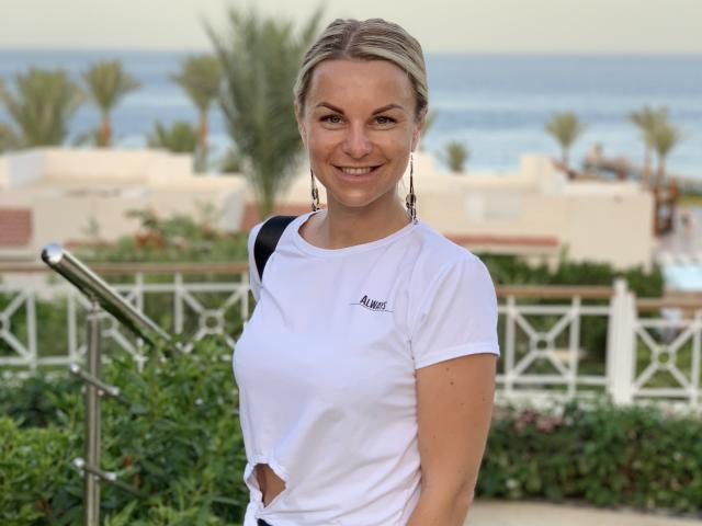 BelleLisaG, a blonde woman with a slight smile, stands in front of a coastal scene. She wears a white, short-sleeve, athletic shirt with a small cutout on the side and "Aimee" logo. Her hair is tied back, and she has long, dangling earrings. The background includes blurred palm trees, white buildings, and a clear blue ocean. She carries a black strap on her right shoulder. The composition focuses on BelleLisaG, with a green plant and white fence in the foreground. The image is brightly lit with natural sunlight.