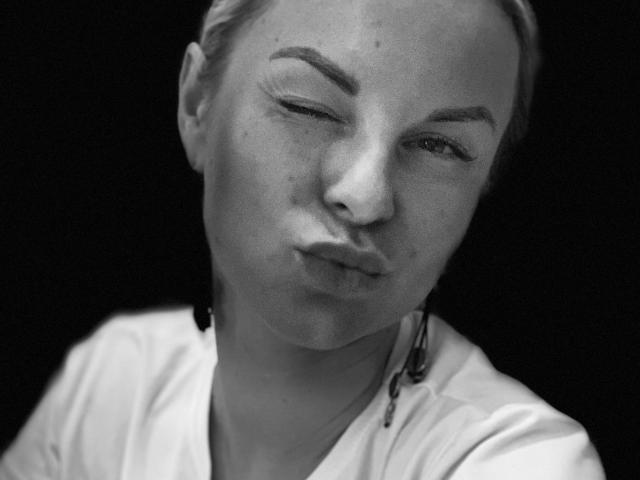 Black-and-white portrait of BelleLisaG with one eye closed and lips puckered. She has light skin, blonde hair pulled back, and visible freckles. Her right eyebrow is slightly raised. She wears a white shirt with a black cord hanging from her right ear. The background is completely black, emphasizing her face. The image has a sharp, high-contrast look with detailed textures on her skin and hair. Her expression is playful and slightly mischievous.