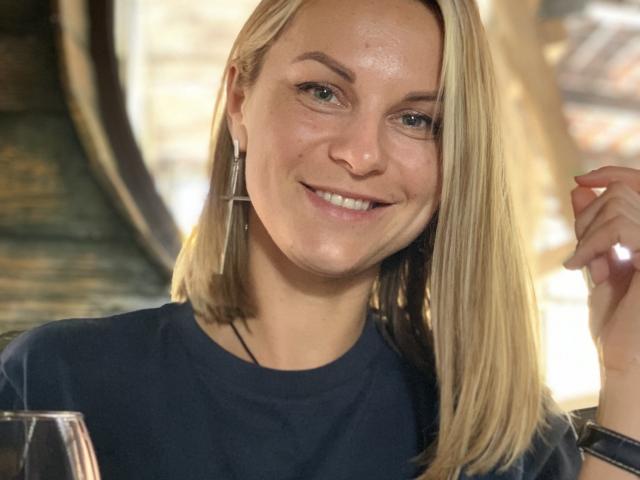 Close-up of BelleLisaG with blonde hair, blue eyes, and a slight smile. She wears a dark blue shirt, thin silver earrings, and a black smartwatch on her right wrist. Her skin is light and slightly shiny. She's holding a glass with her right hand. Soft, natural light illuminates her face. The background is blurred. Her hair is straight and parted to the side.