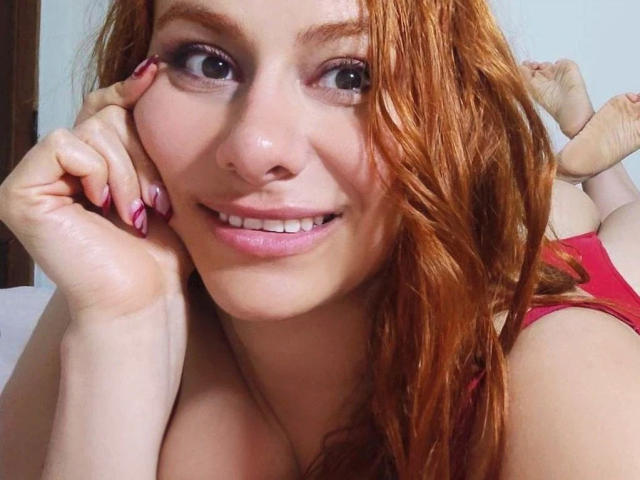 GeileLorena-hot with red hair, pink nail polish, and red lingerie, smiling at the camera, her hand resting on her cheek, showing her tits and cleavage.