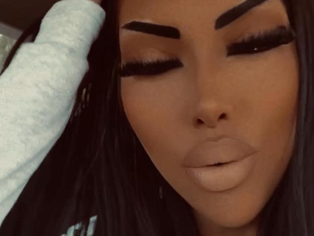 LuxuryDoll69's face with dark brown skin, thick black eyebrows, long eyelashes, and full, glossy lips. A hand in a white glove touches her head.