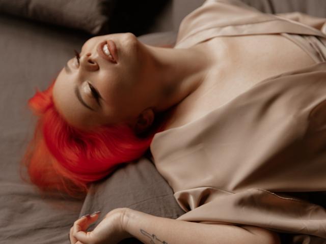 EnergyBurnny with bright red hair, lying on a bed, smiling with eyes closed, wearing a silky beige top, arms relaxed, small tattoo on wrist, soft light, relaxed vibe.