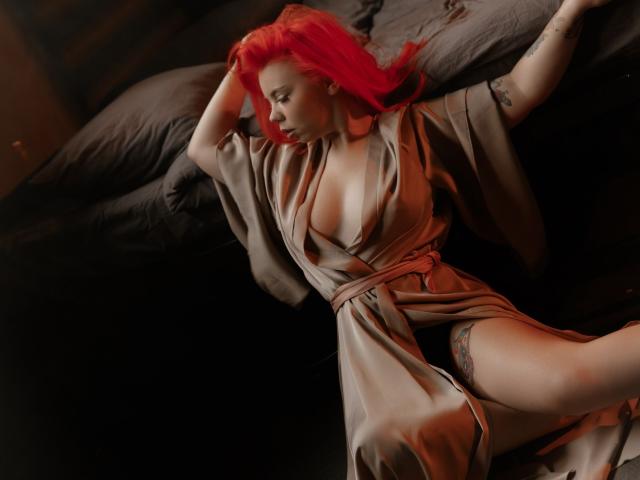 EnergyBurnny with bright red hair, lying on a bed in a beige satin robe, exposing her tits, thigh tattoo, and a hint of her pussy, arms raised, looking down.