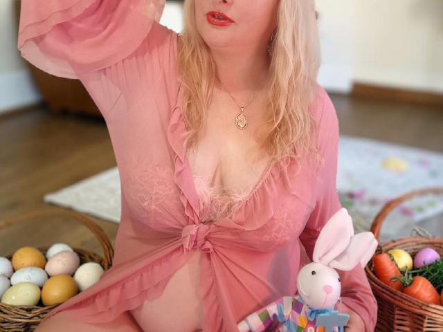 KelliRedhaer in pink sheer robe, showing her tits, with blonde hair, red lipstick, and a bunny toy in her lap, surrounded by Easter eggs and baskets.
