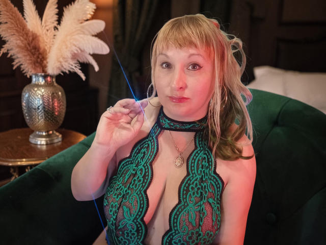 KelliRedhaer sits on a green velvet couch, wearing a green lace halter top that reveals her large breasts. She has blonde hair with bangs and looks at the camera with a slight pout. She holds a blue string taut with her right hand. A silver vase with beige feathers is on the wooden table to her left. Warm lighting casts a red hue on her face. She wears a small gold necklace. The image is a photograph.