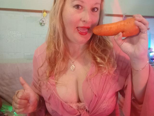 KelliRedhaer with blonde hair, red lipstick, pink dress, and visible cleavage, holding an orange carrot to her tongue, giving a thumbs-up, wearing a necklace.