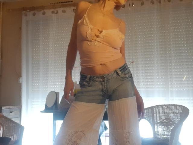 IssahneMay stands in a sunlit room, wearing a pink, lace-trimmed, spaghetti strap top that reveals her small breasts and a slightly protruding belly. She also has on short, blue denim shorts and white, lace-topped leggings. Her head is tilted down, making her face partially visible. The background includes white, semi-transparent curtains and a wicker chair to the right. The lighting is natural, with sunlight streaming through the curtains. The image has a casual, slightly cluttered feel.