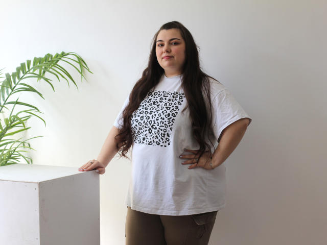 MileyMiln, chubby with long dark hair, wearing a white t-shirt with a leopard print, brown pants, standing with hands on hips, leaning on white cube, green plant on left.