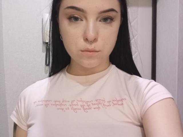 VeronikaGleim with pale skin, black hair, and a nose ring, wearing a white shirt, staring at the camera with a neutral expression, showing her tits slightly.