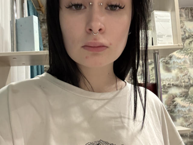 JosetteKarns with pale skin, black hair, nose ring, and three upper lip piercings, wearing a white shirt, looking serious, slight zit on right cheek, in a shelf-filled room.