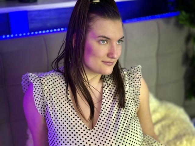 StephieRouse with wet brown hair in a ponytail, wearing a white polka dot dress, smiling slightly, sitting on a gray couch, with purple and blue lighting.
