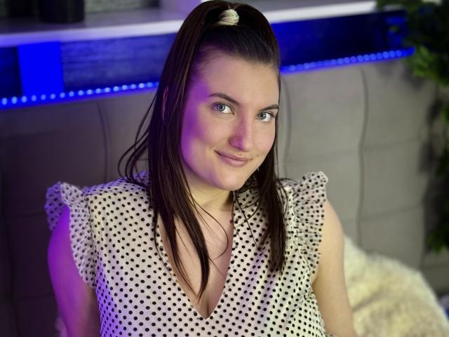 StephieRouse, avec des cheveux bruns attachés en queue de cheval, portant une robe à pois, souriant légèrement, avec une lumière bleu-violet sur sa gauche, et un coussin blanc derrière elle. Ses seins sont visibles..