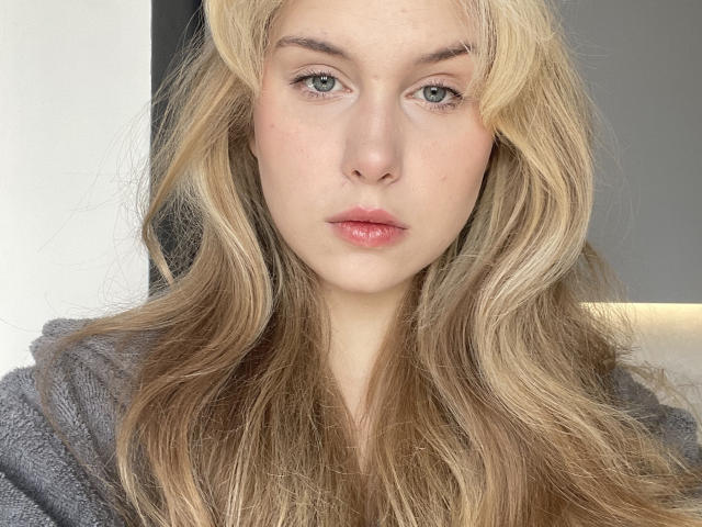 ShenitaMarts with blonde wavy hair, pale skin, green eyes, and pink lips, wearing a grey hoodie, looking straight at the camera with a neutral expression.