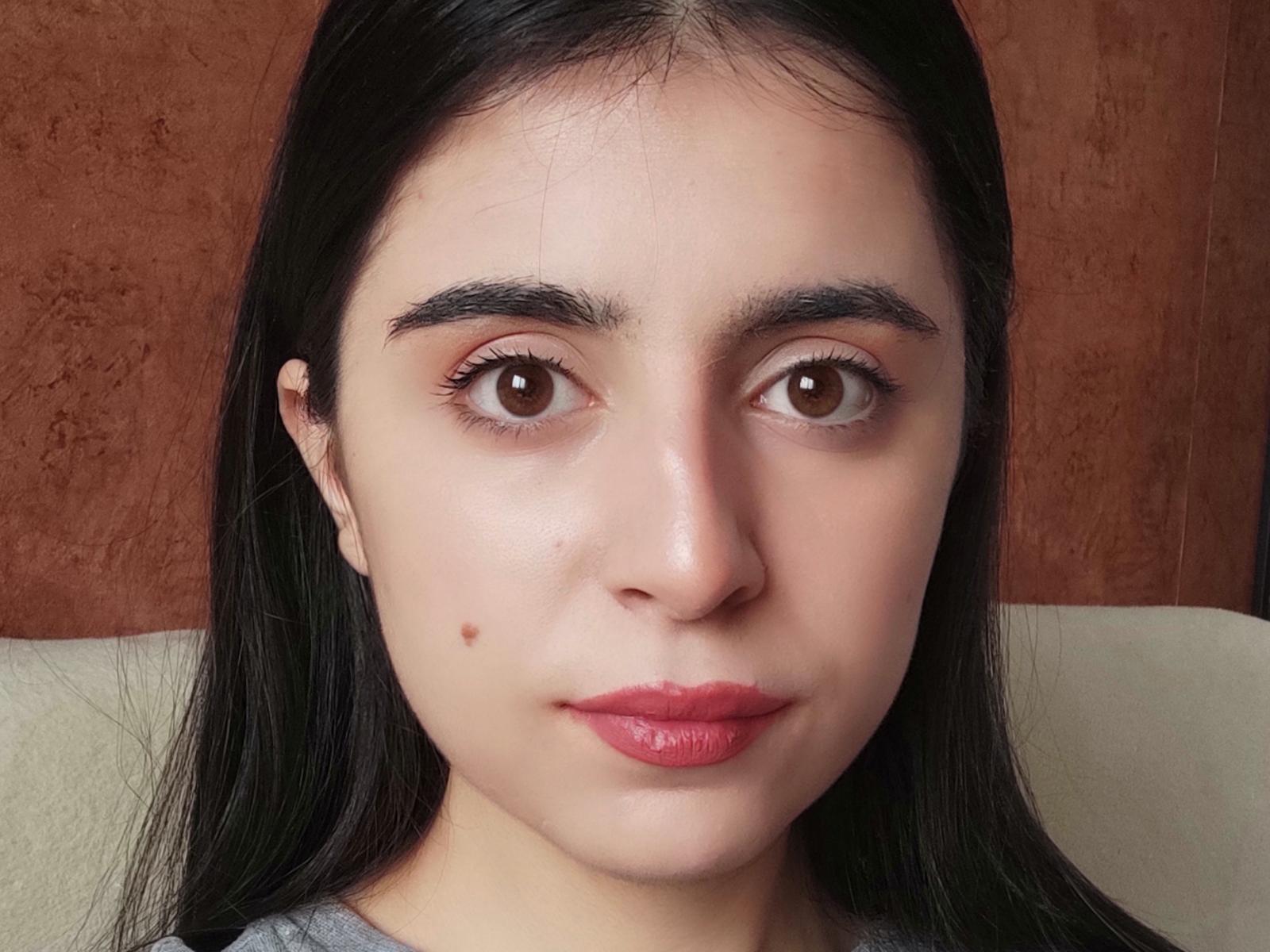 MariaHebdon has brown eyes, black eyebrows, long black hair, pink lipstick, and a small mole on her left cheek. She's wearing a grey shirt.