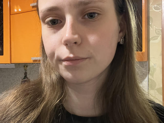 AldaDenham has pale skin, brown eyes, and long brown hair. She's wearing small earrings and a black shirt, with an orange cabinet in the background.