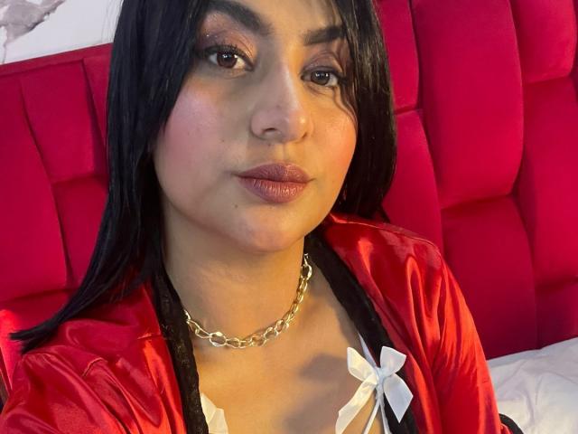 DiannaRosse with black hair, brown eyes, and pink lipstick, wearing a shiny red satin robe with black trim and a gold chain necklace, sitting against a red headboard.