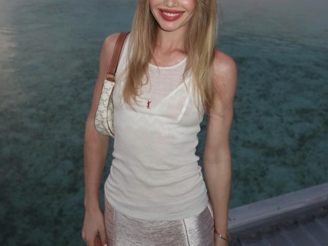 TheHottestBlonde with long blonde hair, smiling, wearing a white tank top, and silver pants, has a small red necklace, and a beige bag on her right shoulder.
