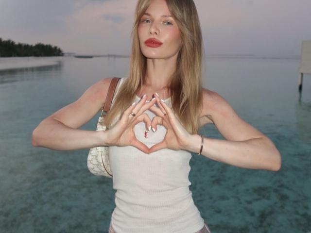 TheHottestBlonde stands in shallow, clear water with a calm horizon in the background. She has long, straight blonde hair, fair skin, and wears a white, sleeveless, ribbed tank top. She holds her hands in a heart shape in front of her chest, with a small red cross in the center of the heart. She has red lipstick and a black hair tie on her right wrist. A small, beige, patterned handbag is on her left shoulder. The image is clear with natural light.