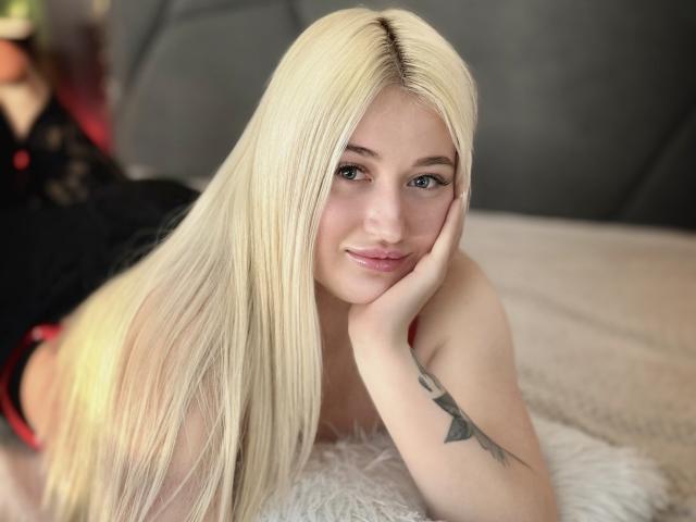 Close-up of HollyTaillor lying on a bed, head resting on her right hand. She has long, straight blonde hair, fair skin, and blue eyes. Her left arm, visible, has a black triangle tattoo on her inner forearm. She's wearing a black top with red trim. The bed has a white, fluffy texture. Her expression is relaxed, with a slight smile. The background is blurred. The image is sharp with a soft focus on her hair and face.