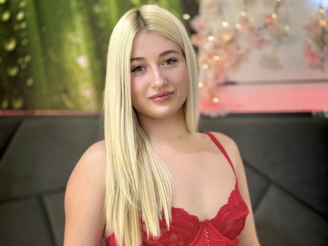 HollyTaillor has straight, platinum blonde hair that falls past her shoulders. She wears a red lace bralette with thin straps. Her skin is fair, and she has a slight, friendly smile. She has light brown eyes and a small nose. The background is blurred, featuring green and pink elements. Her bralette is detailed with lace patterns. The image has a soft, natural light that highlights her hair and skin. The overall composition is centered on HollyTaillor, with a focus on her upper body.