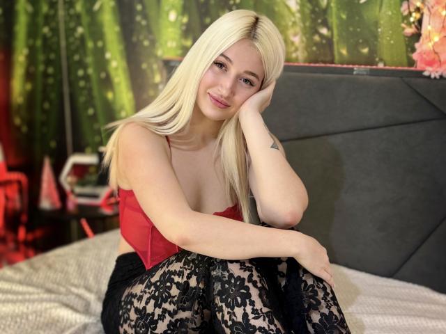 HollyTaillor with blonde hair, red top, black lace skirt, smiling, resting head on hand, showing small tattoo on arm, sitting on bed, looking cute.