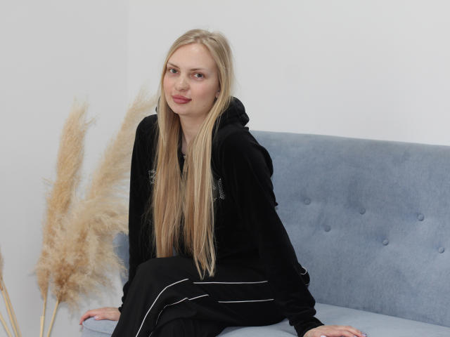 NancyBrooks with long blonde hair, black hoodie, and black pants, sitting on a blue couch, smiling slightly, with puffy beige grasses to her left.