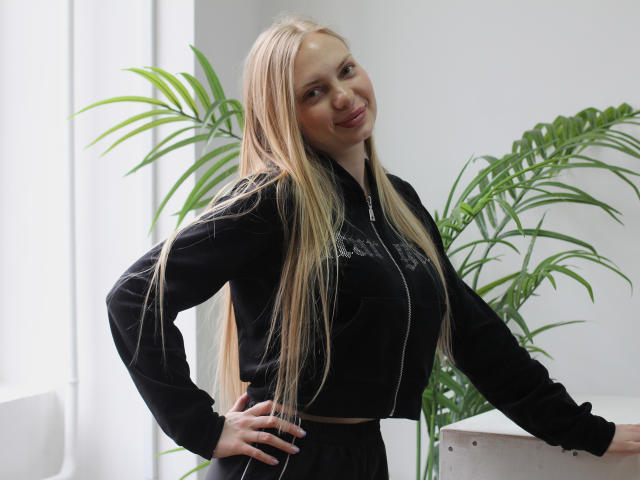 NancyBrooks stands with one hand on her hip, smiling slightly. She has long, straight blonde hair and fair skin. She wears a black zip-up jacket with a white zipper and a black skirt. Her right hand rests on a white surface. A large green plant with long, thin leaves is in the background. The image uses natural lighting. Her face is slightly turned to the right, and she has a confident, relaxed expression. The composition is simple, with a focus on NancyBrooks and the green plant.