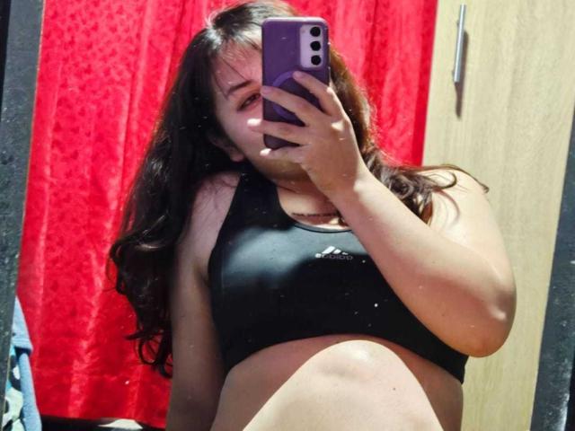 A selfie of MaddieLesYoux shows her holding a purple phone in front of a mirror. She has long, wavy brown hair and is wearing a black sports bra with a small white Adidas logo. Her midriff is visible, and she has a slightly visible belly. The background features a red curtain and a wooden door. The image has a casual, informal style, with slight reflections on the mirror. The overall composition is centered and focuses on MaddieLesYoux's upper body.