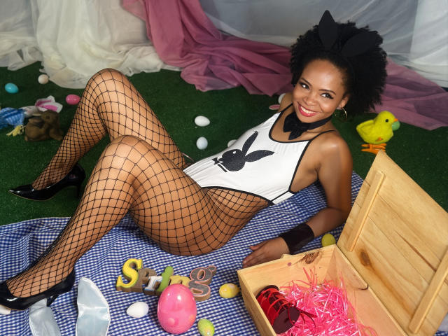 ChantalHaneke, black woman with curly hair, wearing fishnet stockings, white bunny leotard, black heels, lying on blue checkered blanket, wooden box with pink straw, yellow chick on her.
