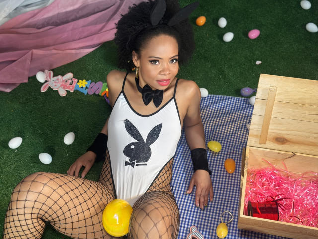 ChantalHaneke, with black bunny ears, fishnet legs, white Playboy top, bowtie, and black wristbands, sitting on blue checkered blanket, Easter eggs, yellow egg in front, wooden box.